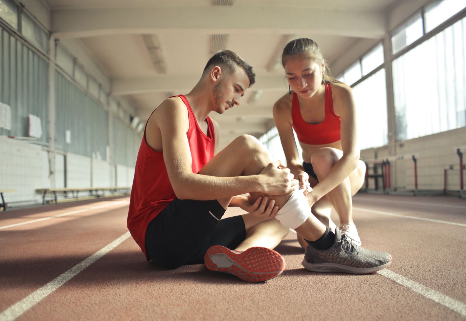 Runner sitting on a running track holding a painful lower leg while a training partner checks on them