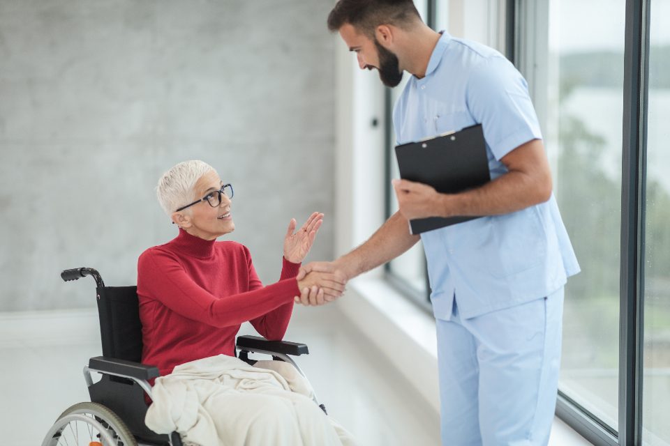 A clinician holding a clipboard greeting an older patient seated in a wheelchair by a window
