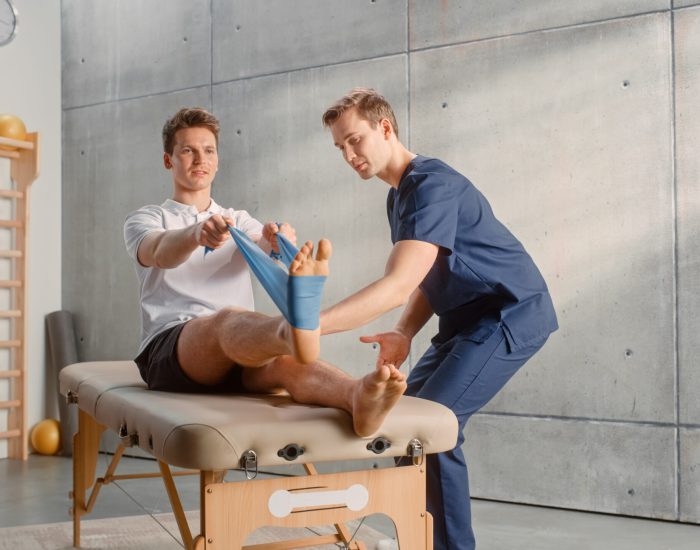 Young Male Athlete Undergoing Physiotherapy, Professional Sport Masseur Helping with Foot Exercise with a Rubber Band. Musculoskeletal Pain Therapy and Rehabilitation in Modern Clinic Concept.